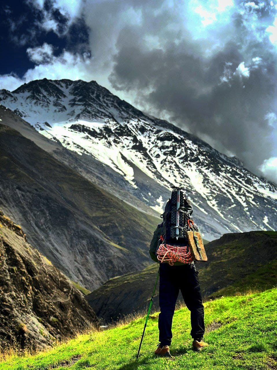 A trekker with a coiled rope and pack on their back, looking up at a steep snow-streaked face under heavy cloud